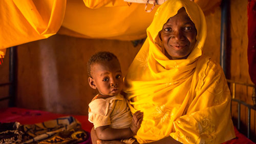 Nana holds her baby son. She is dressed in a vivid yellow scarf