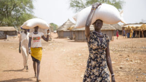 Two tall young women carry large bags of grain on their heads.