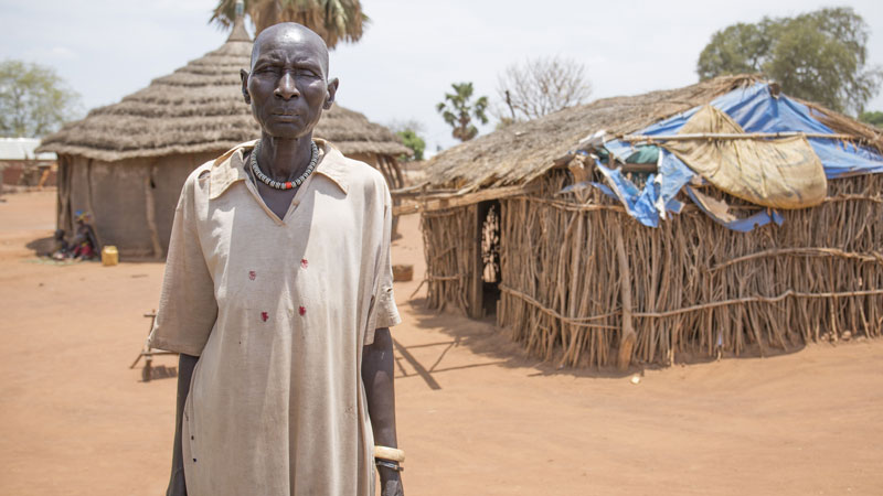 A tall but frail lady, Mary, stands in front of a small building made from branches