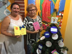 Jenilda and Zeza stand holding Christmas messages beside a Christmas tree in the Cultural Centre in Vila Prudente. There is a CAFOD bauble on the Christmas tree.