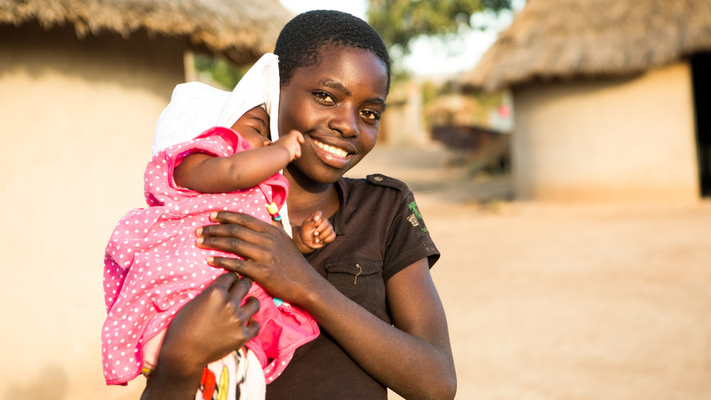 A 12-year-old girl holds her baby sister in Zimbabwe. Their mother, Marian has been supported with vegetable seeds, nutrition and farming training, and fenced off a community vegetable garden to keep it safe from livestock. Marian is now growing a range of nutritious vegetables, nuts and beans for her children