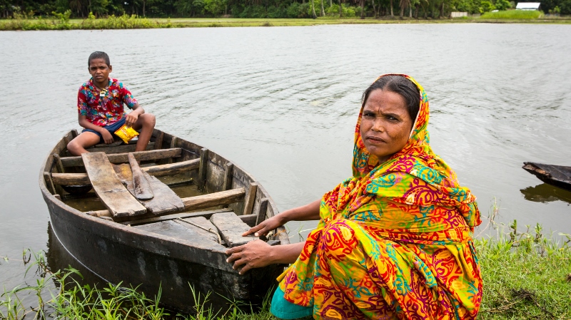 Mahinur crouches by the riverbank holding a small boat with her son Rabiul in,