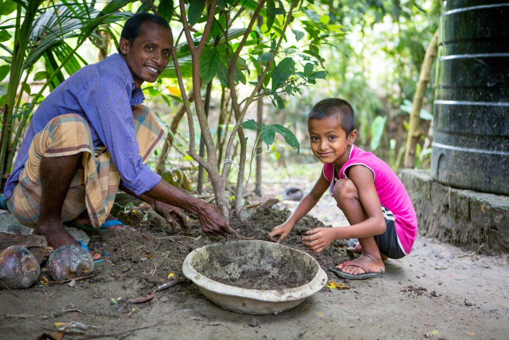 A man and a small boy smiling and digging in the mud in their garden in Bangladesh