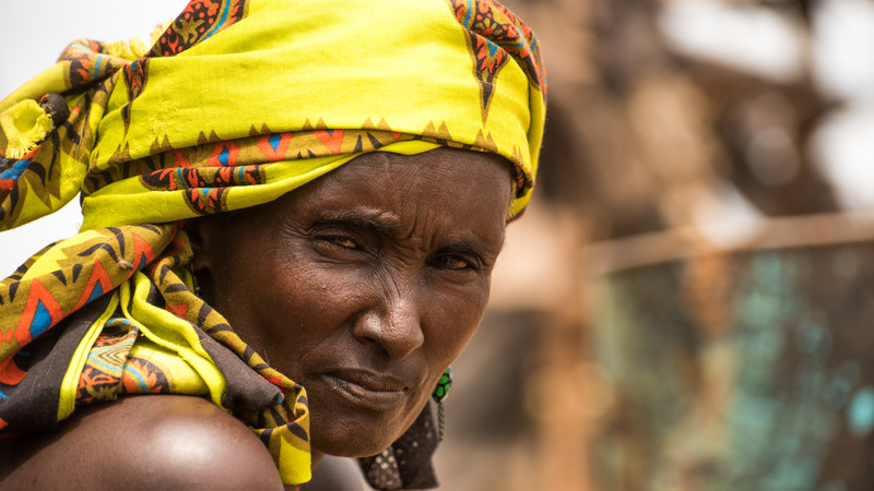 A woman wearing a yellow headscarf looks directly at the camera turning her head slightly to the right