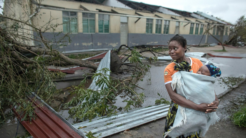 A woman holding her baby standing next to a building wwhose roof has been destroyed in Cyclone Idai in Mozambique. There are trees and sheets of corrugated metal on the floor at her feet