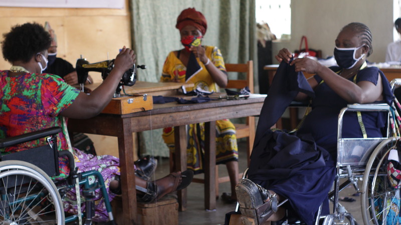 A group of women wearing masks sitting around a table working on making more masks. One woman, in a wheelchair, uses a sewing machine. The others fold material or hold up the finished object