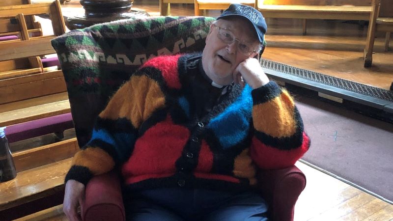 A priest wearing a woolen cardigan sits comfortably in a chair