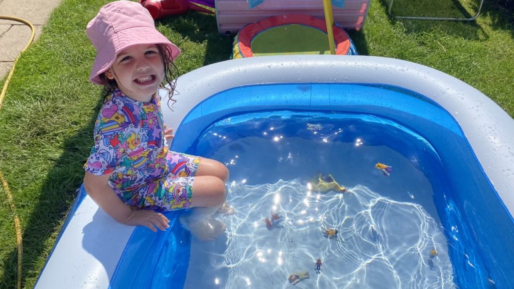 Pippa sits on the side of a paddling pool. There are small toys floating in the water