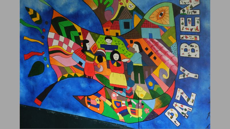Painted mural of a dove filled with images of people and places, next to the words 'Paz y bien.'