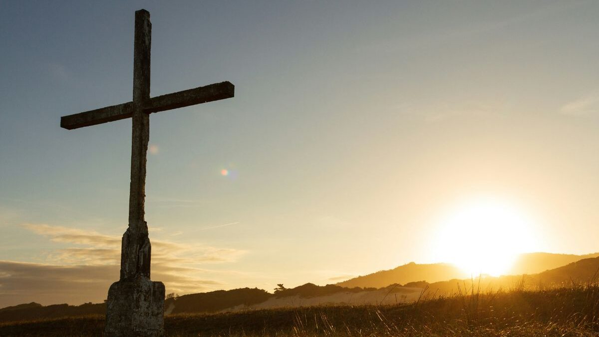 A large cross in a field in the foreground, with a the sun setting behind.